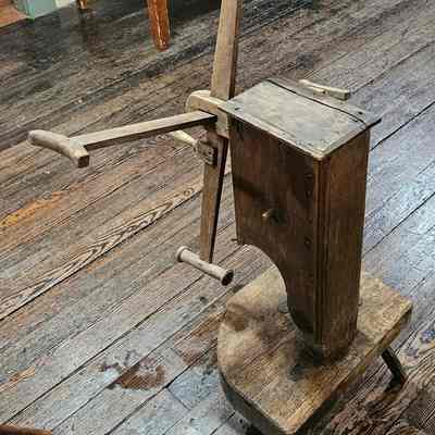Yarn Winder, Back and Side View.: The image depicts an old wooden yarn winder or click reel and crank apparatus, sitting on a worn hardwood floor. The winder appears to be handcrafted, featuring a rectangular wooden body with a handle protruding from one side, used for rotating the internal arms. The floorboards are dark and show signs of age and wear, adding a rustic, historical element to the scene. In the background, the legs of a table and additional chairs are partially visible, hinting at a traditional or vintage setting, perhaps in a historical kitchen or museum exhibit showcasing antique equipment.