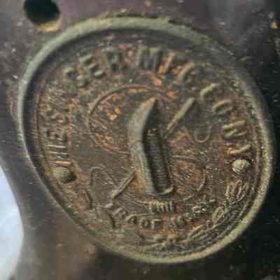 Sewing Machine, Close Up Showing the Singer Logo/Badge. Before Cleaning.: The image shows a close-up of a vintage metal emblem featuring the words "SINGER MFG. CO." and "MADE IN USA" encircling a central design that resembles a screw or a bolt with a letter "S" prominently displayed; the emblem appears to have a patina, suggesting age, and the overall circular motif has intricate detailing around the edges, giving it a distinct antique appearance that is enhanced by the texture of the metal, which shows signs of wear and oxidation, hinting at its historical significance or use in an industrial context, invoking a sense of nostalgia or appreciation for craftsmanship from an earlier era.