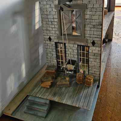 Left Side View: The image depicts a detailed miniature model of a rustic building with a weathered stone facade and a small wooden platform in the front. An intricate pulley system with a spool of rope is mounted above a large window that has two doors open, revealing the inside of the structure. The platform is adorned with several small wooden crates and two bundles wrapped in twine, suggesting a historical or industrial setting. The craftsmanship of the model highlights an attention to detail, with carefully constructed steps leading up to the platform and a realistic texture on the walls and flooring. The entire scene is set on a wooden base, placed on a wooden surface, which enhances the authentic appearance of the miniature structure.