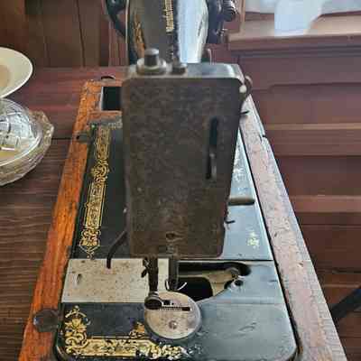Singer Sewing Machine, Side View: The image shows an antique sewing machine with a beautifully crafted metal body, displaying intricate designs, and mounted on a wooden base with elaborate decorations, sitting in a room with a wooden table, a plate, a glass bowl, a framed artwork on the wall, and a softly lit window with white curtains, creating a nostalgic and vintage atmosphere.