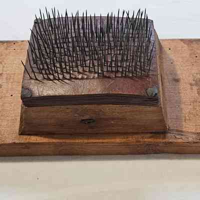 Fine Hetchel: The image shows a hetchel -- a wooden board with a rectangular section featuring numerous sharp, thin metal spikes protruding vertically from its surface. The board appears aged, with a weathered texture and several small holes scattered across its surface. A small label with writing is attached to the side of the board with a string, possibly indicating an item number or classification. The overall appearance suggests this object might be a tool or contraption, perhaps used historically for activities like working flaxl, where fibers are separated and aligned.
