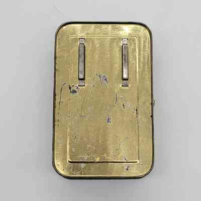 The image shows a vintage, rectangular, metal container with visible wear and two parallel, silver clasps