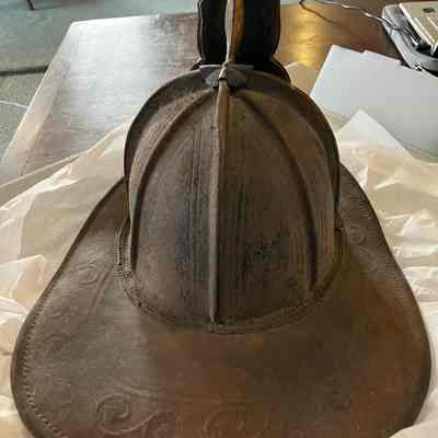 Back view of fire helmet: This image shows a view of the helmet from the back side.  It is made primarily out of brown leather. There is some fading of the brown leather on the back showing some black underneath. There is a clip on the back of the helmet with a metal loop. At the top of the hat is a metal eagle clipping onto the front. There is a decorative imprint in the leather around the rim of the hat.
