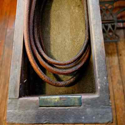 Close Up of Sewing Machine Table Drawer, with Coiled Leather Drive Belt. Before Cleaning.: The image shows the interior of an old wooden drawer featuring a coiled belt, probably a part of a vintage sewing machine mechanism. The drawer, characterized by its weathered and aged appearance, displays a rich, dark wood grain texture. The coiled leather or rubber belt inside hints at its function, likely used to connect the machine's treadle to the driving wheel, facilitating manual operation. The drawer appears to be a component of a treadle sewing machine, indicated by the glimpse of the metal treadle pedal visible in the background, known for their historical significance and mechanical ingenuity. The scene is illuminated by soft, natural light, emphasizing the nostalgic and antique appeal of the piece, with the warm tones of the drawer wood contrasting against the rustic wooden floor