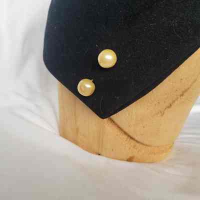 A wooden mannequin head displays a black hat adorned with two pearl buttons against a white background.