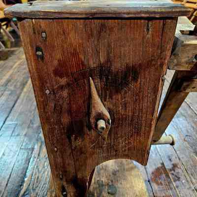 Yan Winder, Showing the Clock and Pointer.: The image depicts an antique wooden yarn winder or click reel sitting on a wooden floor in what appears to be an old-fashioned room or museum setting. The mill is made of dark, weathered wood, showcasing intricate craftsmanship with visible grain and texture. It has a central, vertical handle attached to the side, hinting at its functionality for manual operation. The background is filled with warm-toned, vintage decor, including wooden chairs and an array of furniture, contributing to a rustic and nostalgic atmosphere. The lighting highlights the rich tones of the wood, adding depth and character to the scene.