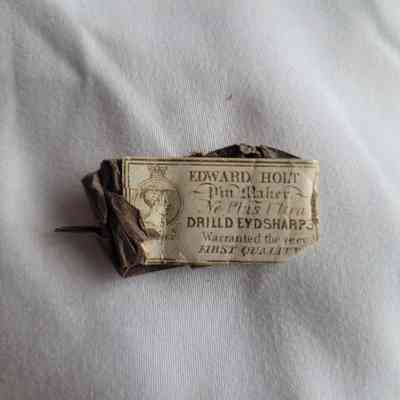 The image depicts an old, crumpled pin label with intricate typography on white fabric.