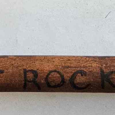 Rest of Inscription on Shaft of Cane: The image shows a close-up of a wooden walking stick with text written on it that reads: "C. WESTERLY R.I. AT ROCKY POINT AUG. 1937." The wood has a natural brown color with visible grain patterns. The text appears to be hand-written onto the surface in black ink, suggesting it may be a souvenir or a commemorative piece marking an event or visit from August 1937 at Rocky Point in Westerly, Rhode Island. The background is a plain, light color, which highlights the wood and the letters, emphasizing the historical and nostalgic aspect of the object.