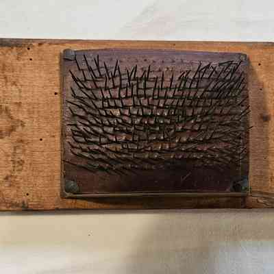 Fine Hetchel, Top View: The image shows an old wooden hetchel, used in traditional textile processing, featuring a rectangular wooden base with a grid of angled, metal spikes protruding from a smaller, raised wooden panel in the center. The wood appears worn and aged, suggesting extensive use over time, with a light brown hue and some darker areas of discoloration. A small tag marked with numbers and letters is attached to one corner using string, indicating it might be part of a collection or inventory. The entire object is placed on a white fabric or surface, emphasizing its rustic and historical character.