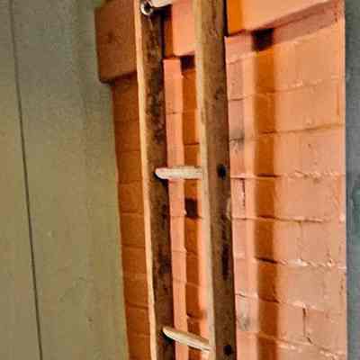 Florimond Bergeron's Ladder.: The image shows a tall, rustic wooden ladder leaning against a textured red brick wall inside what appears to be an indoor space. The ladder's rungs are evenly spaced, and the natural wood grain is visible, adding a classic, aged charm to the setting. The wall features a distinct seam where orange and dark brown meet, likely part of the building's structural or decorative elements. The floor is dark and polished, contrasting with the warm tones of the brick. A small circular hole is visible on the floor.