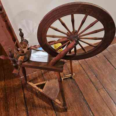Hopkins Flax Spinning Wheel on Display at the Mill Museum.: The image shows a wooden spinning wheel, a classic tool used historically for spinning fibers into thread or yarn. It features a large wheel with spokes connected to a smaller axle and a traditional treadle foot pedal below, all mounted on a sturdy wooden frame. The spinning wheel has a rich, polished finish, reflecting the craftsmanship of its maker. It stands on a wooden floor and is positioned against a white wall. Attached to the spinning wheel is a piece of paper, which might provide further information about its history or details of its construction. Overall, the spinning wheel evokes a sense of nostalgia and serves as a reminder of the traditional methods of textile production in earlier times.