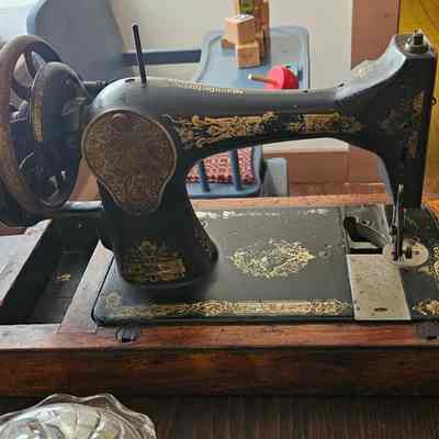 Singer Sewing Machine, Back View: The image depicts a vintage sewing machine with ornate gold detailing, set within a wooden base, positioned on a wooden table next to a glass dish; in the background, there are some blocks and a toy resting on a blue chair, and soft daylight streams through a window, casting gentle shadows across the scene, evoking a sense of nostalgia and craftsmanship in a cozy, homey environment that feels reminiscent of a bygone era.