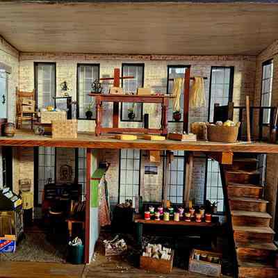 Interior View, Showing All Three Rooms: The image depicts a detailed miniature model of a two-story interior thread mill. The upper level features a wooden table with crafting tools, surrounded by various items like a wicker basket, a chair, and barrels. There are decorative elements including small sculptures and dried flowers hanging from racks. On the lower level, there's a desk with a vintage cash register, books, and a bulletin board. The workspace includes a variety of tools, colorful spools of thread or paint, and other crafting materials. A staircase connects the two levels, and its walls are adorned with large windows and a calendar, adding an authentic touch to the scene.