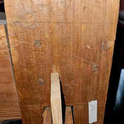 Wooden pegs. (The wedge is new.): The image showcases a close-up view of a wooden loom, highlighting the texture and grain of the wood. The wood appears worn, indicating age, with visible wooden pegs and small marks scattered across its surface. At the center, there is a protruding wooden piece, likely a joint or peg, which joins with another section, emphasizing a traditional carpentry technique. The wood has a warm, natural color, and a small white sticker with the number "3" is affixed to the lower right side, suggesting it might be labeled for identification or categorization purposes. The setting is dimly lit, casting soft shadows that accentuate the wood's aged character and natural imperfections, giving the piece an aura of historical character.