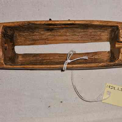 Throw Shuttle: The image shows a wooden throw shuttle with a long, narrow, and rounded shape, featuring a central rectangular cutout that suggests it might be part of a traditional weaving or textile-making process. The wood appears aged and weathered, adding texture and character to its overall appearance. Attached to the object with a piece of string is a small yellow tag labeled "1958.L.270," suggesting this item might belong to a collection or has been cataloged for inventory or display purposes. The background is a plain, light-colored fabric that contrasts with the wooden object, drawing focus to its details and contours.