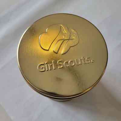 A gold metallic tin lid displays the Girl Scouts logo on a light background.