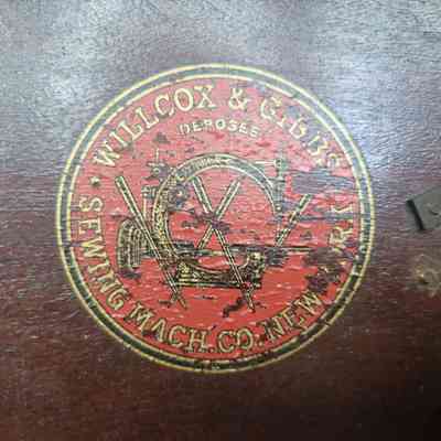The image shows the logo of Willcox & Gibbs Sewing Machine Company on a wooden surface.
