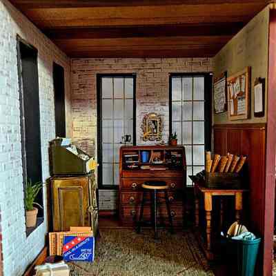 Interior View, Mill Office: The image depicts a quaint, vintage-inspired office characterized by its brick walls and wooden ceiling. Two tall windows are present in the back, allowing natural light to filter into the cozy space. In the center is a wooden desk with an assortment of items, including books and stationery neatly arranged, beside it stands a stool. A small potted plant sits atop the desk as a decorative touch. To the left is a vintage green cabinet, which holds various papers and office supplies, and a couple of packages are placed on the floor nearby. The right side of the room features a wooden table with stacked papers and folders, while the wall hosts a bulletin board filled with notes and a calendar. The overall ambiance is rustic and organized.