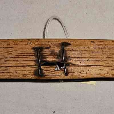Throw Shuttle, Side View, Showing Repair: The image shows a long, narrow wooden throw shuttle with a curved, boat-like shape. The wood has a weathered appearance, with visible texture and grain, suggesting age and frequent use. At the center, there is a metal piece fastened into the wood. The object lies on a plain, light-colored fabric background, emphasizing its rustic and utilitarian design. The overall appearance suggests it could be an antique or an artifact with historical significance, reflecting cultural or regional craftsmanship.