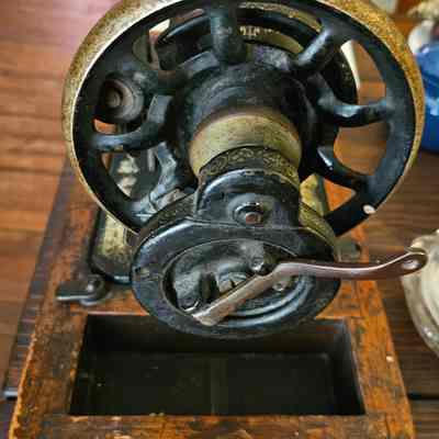 Singer Sewing Machine, Crank Wheel: The image depicts a vintage sewing machine, emphasizing its intricately designed hand-crank mechanism and beautifully aged metallic parts. The machine is set on a wooden base, showcasing its rustic charm with visible wear and patina, suggesting a long history of use. The wheel and handle are prominently featured, indicating the manual operation typical of older models. In the background, a wooden surface complements the antique aesthetic, while other objects add context to the piece's timeless appeal. Together, these elements evoke a sense of nostalgia