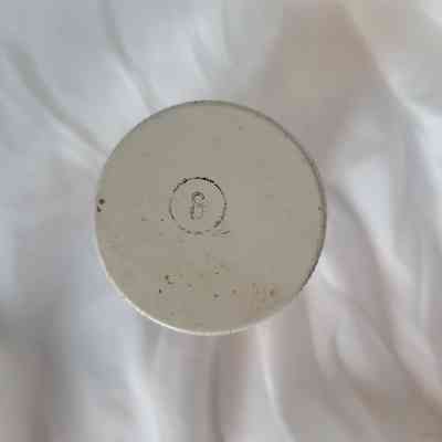 The image shows a white jar lid with a small symbol on top, lying on a textured white surface