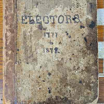 Electors, 1777 to 1878.: The image shows an aged, worn-out book with a leather-bound spine, titled "ELECTORS" and covering the years "1771 to 1878," resting on a wooden surface. The book's cover displays visible signs of wear and discoloration, suggesting it has been well-used over time. There are various dark stains and blemishes on the surface, enhancing its antique appearance. Additionally, a ruler is placed at the bottom of the image, indicating the book's size in inches.
