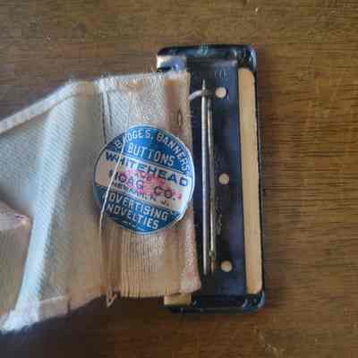 The back of the pin, featuring a round manufacturer's sticker stuck to the ribbon fabric. The sticker is blue on the top and bottom with a band of white in the middle. There is white and blue writing on the respective sections and a red oblong stamp in the middle with different writing.