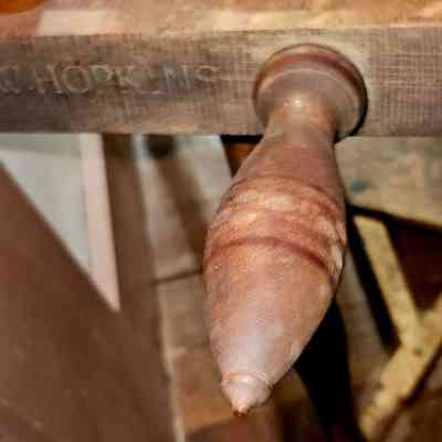 Inscription Carved on Hopkins Flax Spinning Wheel.: The image displays a close-up view of a wooden handle attached to a spinning wheel. The handle is crafted from smooth, polished wood, exhibiting a rich, natural grain pattern. The end of the handle tapers to a rounded point, suggesting it is designed for easy gripping and turning. The wheel has an engraved marking that reads "W. HOPKINS," indicating the maker. The texture of the wheel is rustic, with visible wood grain and a weathered appearance. In the background, a wooden stool or chair is partially visible, with crossed legs adding to the workshop setting. The lighting in the image casts a warm tone, highlighting the craftsmanship and detail in the wood.