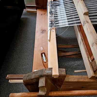 New front beam and bench.: The image depicts a traditional wooden weaving loom, showcasing a partially completed black and white textile design in progress. The loom has a sturdy wooden frame and features various components such as beams and tension rods, all crafted from a rustic, natural wood material. A wooden shuttle, holding a spindle of yarn, is positioned on the loom’s surface, suggesting ongoing weaving activity. The loom is set on a carpeted floor, indicating an indoor setting, perhaps in a museum exhibit room. Its detailed craftsmanship and arrangement highlight the intricate nature of traditional weaving techniques and the artisan's skill involved in creating woven textiles.