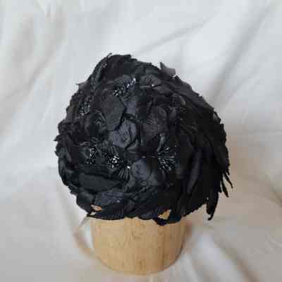 The image shows a black, floral-textured hat sitting on a wooden stand against a white background.