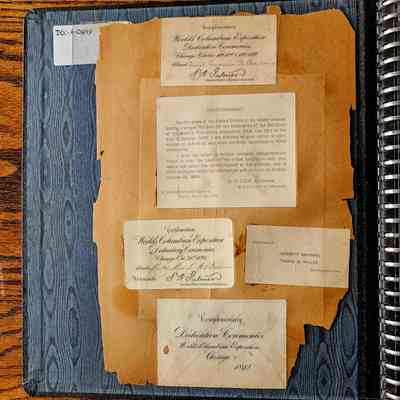 Boss's Scrapbook, Page 2: The image shows an old scrapbook page with several pasted cards and documents related to the World's Columbian Exposition Dedication Ceremonies from 1892. The background is a wooden table, and the page is affixed inside a spiral-bound book. One document details a postponement of the ceremonies, attributed to a congressional decision, and specifies a new date in October 1892. There are "Complimentary" tickets intended for specific guests, signed by authoritative figures from the exposition. The cards are aged, with some discoloration and fraying at the edges, emphasizing their historical significance. The page also includes a label at the top left with the code "DOC-A-0849," possibly cataloging this specific page or item.