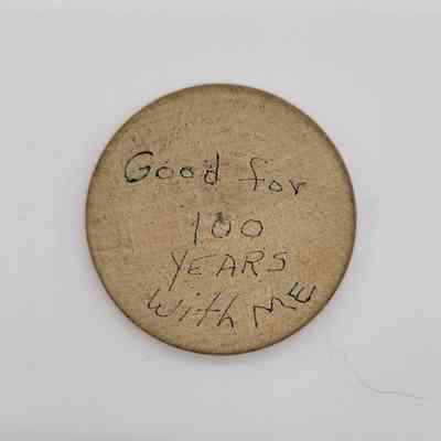 The image shows a round, wooden token with handwritten text saying, "Good for 100 years with me"