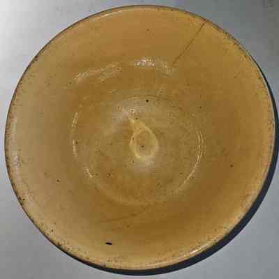 Yellowware Mixing Bowl. Top View.: The image shows a simple, round, beige ceramic bowl placed on a flat surface with a plain background. The bowl appears to have a slightly uneven texture with a few visible specks and a subtle shine, suggesting it might be glazed. There is a noticeable crack near the edge of the bowl, indicating some wear or age. The interior surface forms a gentle curve leading to a smooth and slightly indented center. Shadowing on the right side suggests overhead lighting, highlighting the bowl’s surface and creating a play between light and dark areas. This minimalistic composition emphasizes the bowl’s utilitarian nature and the subtle imperfections that lend it character. The earthy tones and understated design give the object a rustic or vintage appearance, perhaps implying it has been used for practical purposes as opposed to decoration.