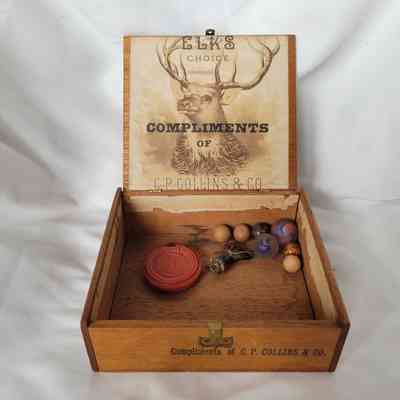 An open wooden box contains marbles, a ceramic boot, and a circular red gutta percha case