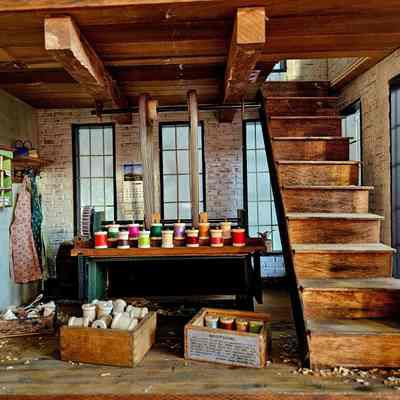 Interor View, Winding Room: The image depicts a miniature workshop room featuring a wooden interior with exposed beams and brick walls, bathed in natural light streaming through large windows. A staircase ascends on the right side of the room, while a winding machine in the center is laden with a vibrant array of colored spools, threads, and various crafting tools. Shelves along the walls are stocked with additional supplies, including a set of draped aprons and neatly stacked, colorful boxes. The floor is scattered with wood shavings, adding to the authentic, rustic charm. A small calendar hangs on one wall, providing a hint of everyday life and routine within this quaint and meticulously detailed workspace.
