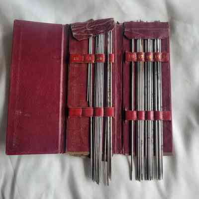 A worn red leather case holds thin steel knitting pins, numbered for organization, against a light fabric backdrop.