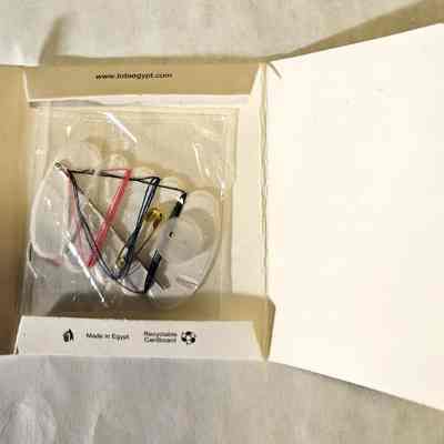 Sewing kit. Open.: The white cardboard container is open, revealing the contents: a celophane bag containing a white button, a small brass safety pin, a stainless steel sewing needle, and four lengths of thread wound onto an oblong holder. The threads are white, pink, blue, and black.