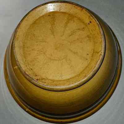 Yellowware Mixing Bowl. Bottom View.: The image depicts an upside-down, circular ceramic bowl with a smooth, yellow-brown finish, slightly weathered with faint marks and spots on its base. The surface reflects a bit of light, indicating it has a glossy sheen. The broad rim of the base is well-defined, and the bowl appears to be set against a neutral, light-colored background, suggesting it might be placed on a table or countertop. The overall appearance of the bowl gives it a rustic and vintage quality, with visible textural details and subtle color variations that add to its character and charm, suggesting it may have been handmade or well-used over time. The shadowing around the bowl's bottom edge emphasizes its round shape and the depth of its basin, which is not visible.