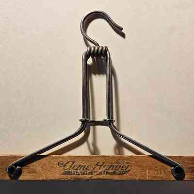 Clothes Hanger for Trousers, Side 2: The image showcases a vintage clothing hanger, featuring a design with a wooden bar at the bottom and a sturdy metal hook at the top, coiled in a unique manner. The metal extends downwards to connect to the wooden part, which bears the text "Acme Hanger, Made in U.S.A." printed on it, emphasizing a classic, durable aesthetic. The background is a plain, neutral tone, which highlights the intricate details of the hanger's construction, showing both functionality and a sense of nostalgia indicative of earlier manufacturing styles.