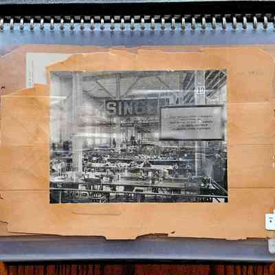 Boss's Scrapbook, p. 1: The image shows an old black-and-white photograph mounted on a worn, brown paper background inside a spiral-bound plastic cover, depicting the interior of Machinery Hall at the Columbian Exposition (Chicago World's Fair) in 1893, with rows of sewing machines and several workers. The photograph focuses on a workspace filled with numerous tables aligned in parallel, each equipped with a sewing machine, emphasizing the extensive scale of manufacturing activity. Above the work area, a sign reading "SINGER" is visible. To the right, a clear sign states, “The Willimantic Company Manufactures the Best Cotton Thread for Shoes ... Exclusively ... School Shoe Factory.” The surrounding plastic cover and the mounting paper suggest that the photograph is preserved for archival purposes.