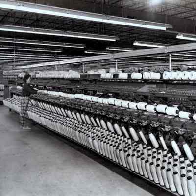 Helen Brown operates a winding machine: A worker named Helen Brown stands in front of a large Foster winding machine in 1966, used for winding manufactured thread onto bobbins. Other winders are in the background. The room stretches to the edge of the frame of the photograph.