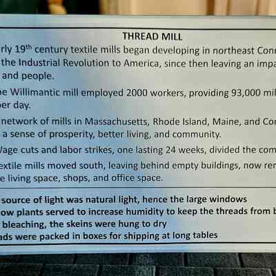 Exhibit Label: The image contains a text about the historical development of textile mills in northeastern Connecticut during the early 19th century, marking the start of the Industrial Revolution in America. It highlights key events: in 1892, the Willimantic mill employed 2,000 workers, producing 93,000 miles of thread daily; by 1898, a network of mills across New England fostered prosperity and community. However, in 1925, wage cuts led to labor strikes, disrupting society. By 1985, mills had moved south, and the buildings were repurposed for residential and commercial use. Additional notes mention natural lighting, window plants for humidity, drying methods, and packaging processes.