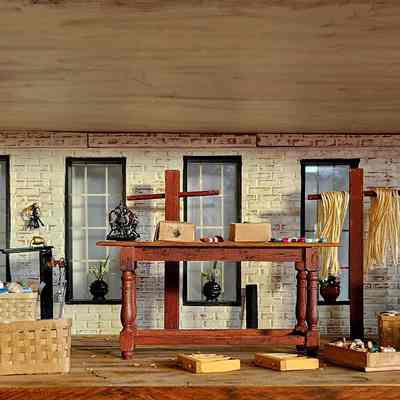 Interior View, Dye Room: The image depicts a detailed miniature room with white brick walls, large windows, and a wooden ceiling that evokes a vintage, rustic ambiance. The room is filled with various textile dyeing materials and tools, such as baskets full of colorful yarn, wooden boxes, spools of thread, and neatly arranged skeins of yarn hanging from a wooden frame. A central wooden table displays more items, while plush plants in vases add a touch of greenery. In one corner, a wooden chair sits beside a pair of barrels.