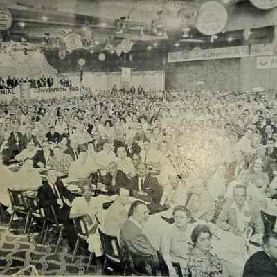 Postcard of Opening Day Ceremonies of 1962 TWUA Convention: The image depicts a large indoor assembly of people attending the Twelfth Biennial Convention in 1962, as indicated by the prominent sign in the background. The room is filled with attendees seated at long tables, creating a bustling and engaged atmosphere. Banners and circular decorations hang from the ceiling, with one visible slogan reading, "Ask not what your Union can do for you but WHAT YOU CAN DO THROUGH YOUR UNION," suggesting a focus on collective action and community involvement. The crowd, a diverse mix of individuals, appears attentive and united in purpose, representing a significant gathering of union members or similar group participants. The ambient lighting and monochromatic tone lend an archival quality to the scene, emphasizing its historical context.