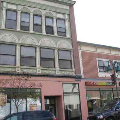 760 Main Street, Windham, CT, from 2024-25 Tax Assessor's Database. (The Building Has Since Been Demolished.): The image depicts an urban street scene featuring a multi-story building with an elaborate facade that includes decorative arches and intricately detailed designs painted in a soft green hue, accented by a pastel base. Adjacent to this structure is another building characterized by a simpler, more modern design, housing a business as indicated by the signage that reads "Copy and Print." In the foreground, a dark-colored vehicle is parked on the street next to a tree, while an American flag is displayed prominently, adding a sense of vibrancy and liveliness to the scene. A streetlamp near the building enhances the urban atmosphere, suggesting a downtown or commercial area where local businesses thrive alongside historical architecture.
