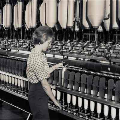 Photo of worker operating spinning machine: An unknown female worker operates a ring spinning machine in 1966. Bobbins of roving (unspun thread) hang from hooks at the top of the machine. Each length of roving is drawn by rollers and then spun into thread, which is wound onto bobbins at the bottom of the machine. The machine handles at least twenty different feeds. The worker lifts a full bobbin of thread off the machine. The worker has short hair. She wears a blouse and what may be trousers.