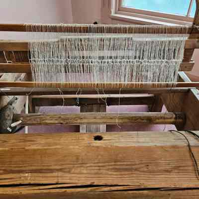 New string heddles. Photo taken before the loom was warped.: The image depicts a vintage wooden loom in a room with pink carpeting and a large window allowing natural light to illuminate the scene. The loom, made of sturdy timbers, is set up with threads intricately arranged, indicating that it is in the process of weaving fabric. The wooden frame includes various components such as heddles, rollers, and beams that are essential for the weaving process. The loom's rustic appearance suggests that it is an old, possibly historical, piece of equipment, used for creating textiles through traditional methods. The room's simple design highlights the loom as the central focus.