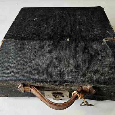 Photo of typewriter case: A photograph of a typewriter case, covered in black faux leather. It has a brown leather handle and silver steel latch. A key is tied to the handle with string.