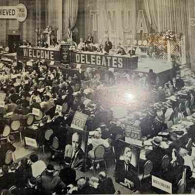 Postcard of Opening Day Ceremonies of 1964 TWUA Convention: The image depicts a large meeting or conference of the Textile Workers Union of America in 1964, with a banner that reads "Welcome Delegates" across the front of a stage adorned with officials seated at a long table. The photograph shows numerous attendees seated in rows beneath signs for different states, such as Rhode Island, Virginia, and North Carolina. Two large portraits of notable individuals are visible amidst the crowd, surrounded by additional signs and placards. The scene reflects the formal and organized nature of a union gathering, where significant business discussions and decisions likely took place. The banner "Much Has Been Achieved" and the years 1939 and 1964 suggest a celebration or reflection on progress made by the union over a 25-year span.