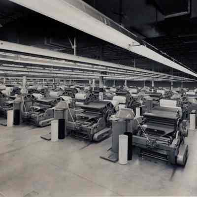 Carding room: A very large room holds dozens of identical large carding machines, used to process raw cotton into sliver in preparation for spinning thread. 1966. The floor appears to be concrete. The absence of overhead drive shafts indicates that the machines are powered by electricity.